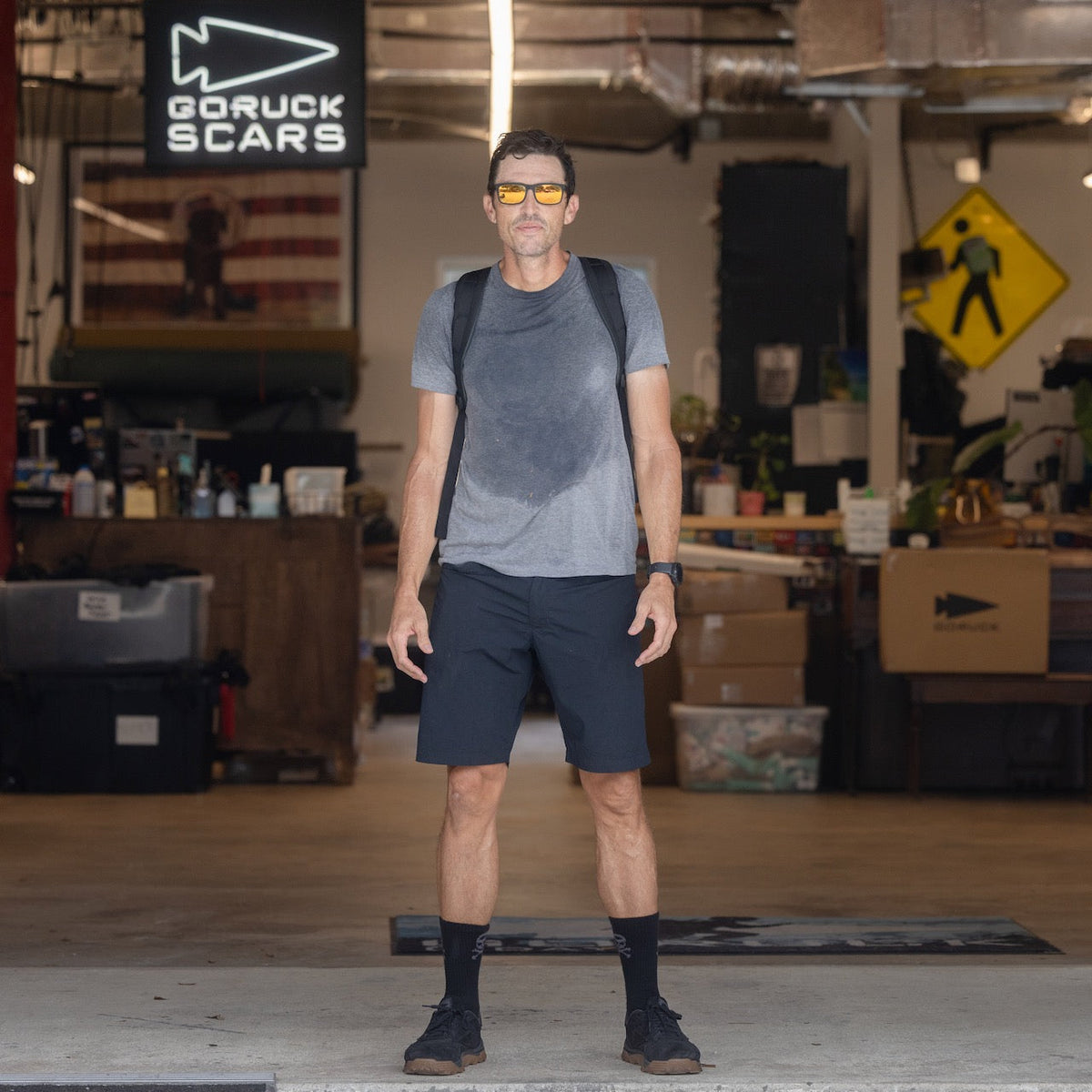A man in a gray shirt, black shorts, and yellow sunglasses stands in an industrial warehouse setting with the Basic Rucker backpack by GORUCK.