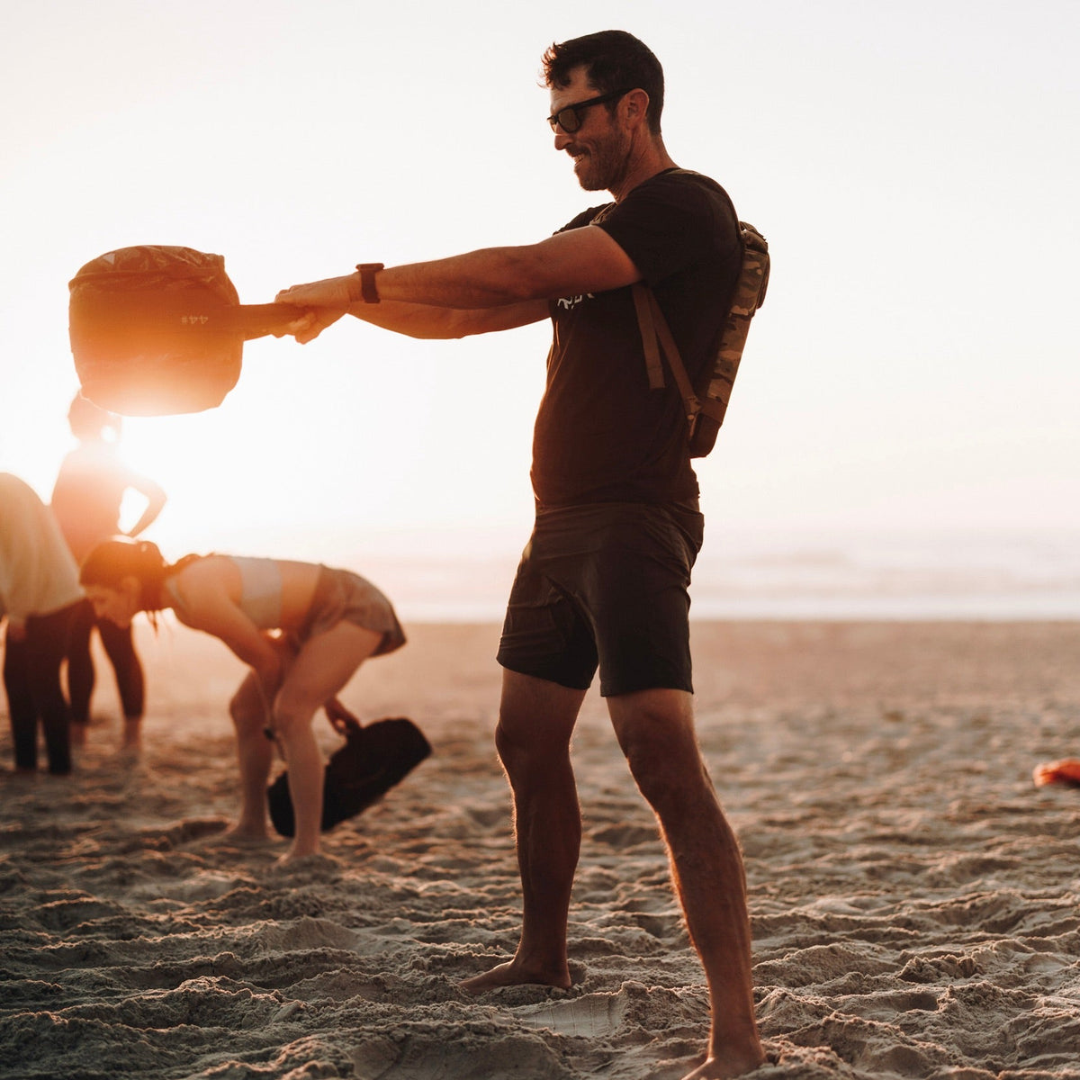 A man exercises with Sand Kettlebells, which feature a heavy-duty padded handle, on a sandy beach at sunset as others work out in the background.