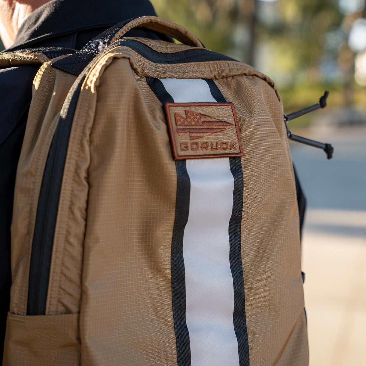 Close-up of a tan Basic Rucker® backpack with a patch and vertical white stripe, worn on someone's back.