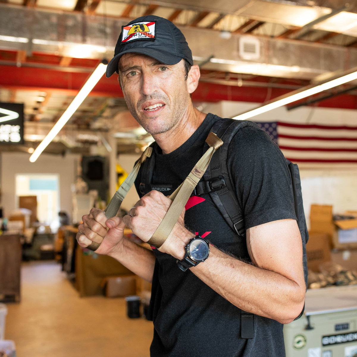A man in a black cap and t-shirt with a backpack stands in a warehouse, American flag behind. T-REX Hand Straps for elbow tendinitis relief peek out from his backpack.