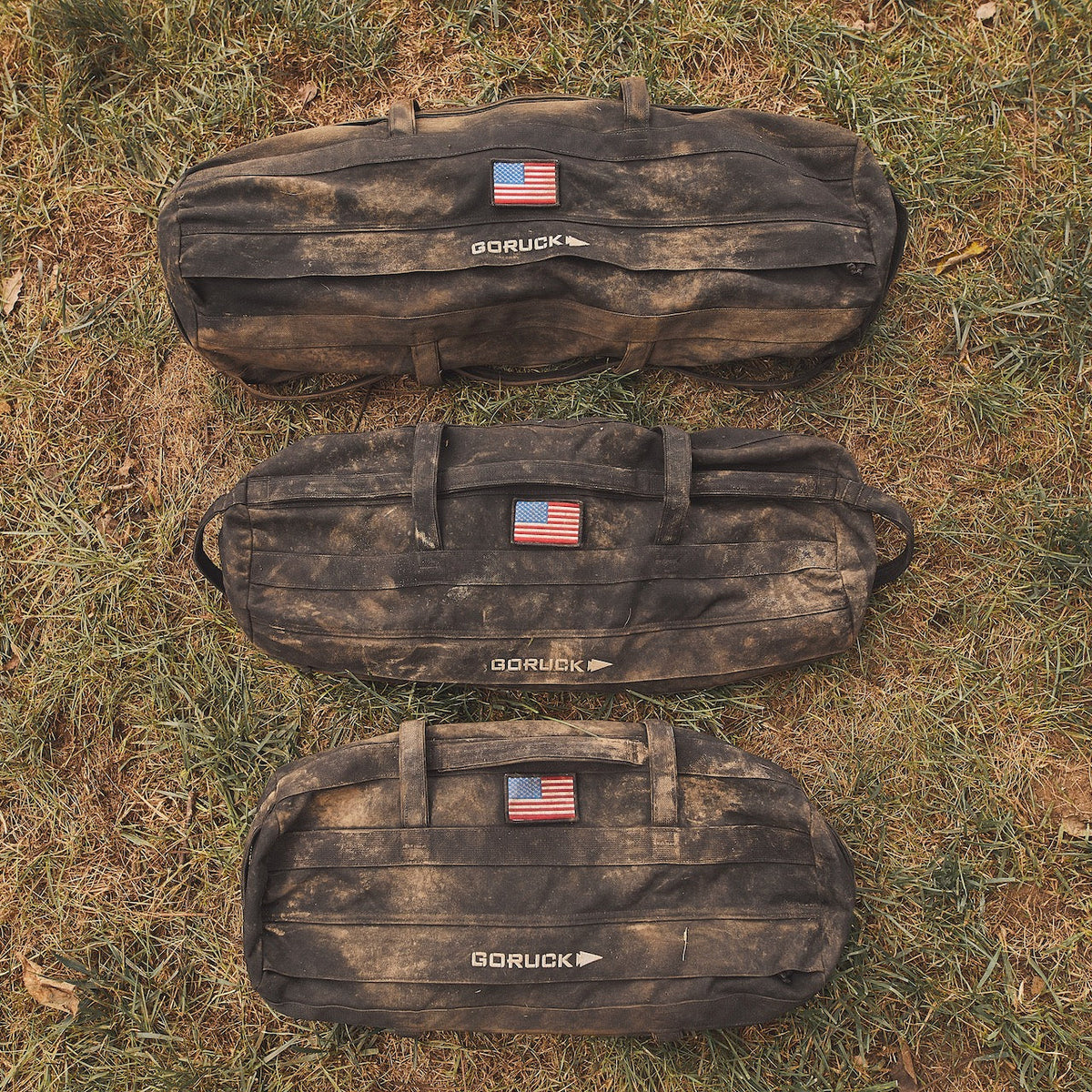 Three rugged Sandbag Kits (w/ Filler Bag), each featuring an American flag patch, rest on the grass—built tough and ready to power your most demanding workouts.