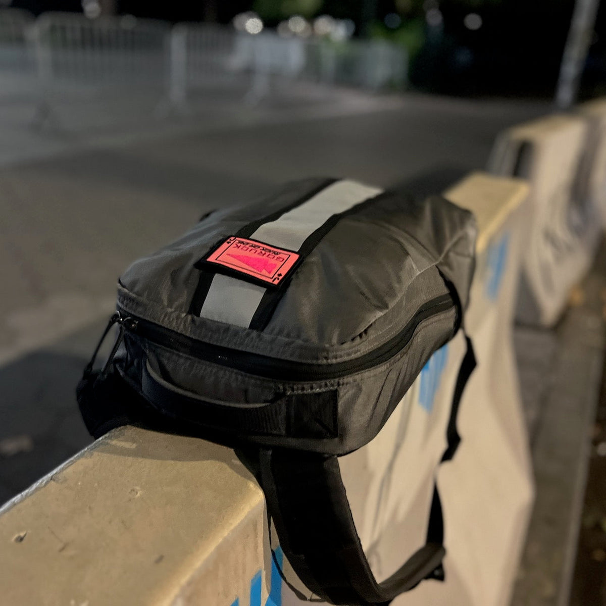 A Basic Rucker in gray, featuring a reflective stripe and a pink label, rests on a concrete barrier at night—ready for your next GORUCK rucking adventure.