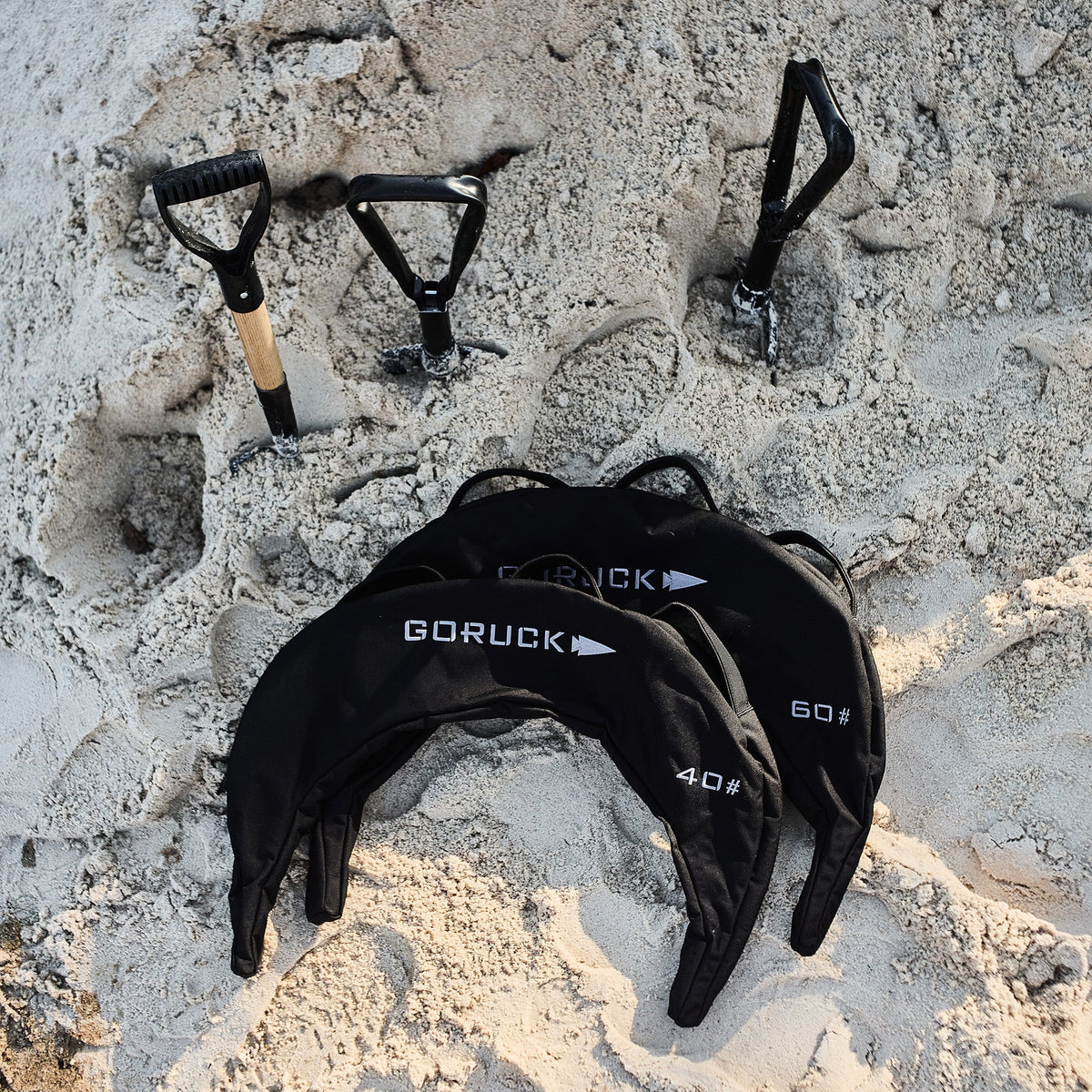 Three shovels and three Bulgarian Sandbags with reinforced handles and weight labels are placed on the sand.
