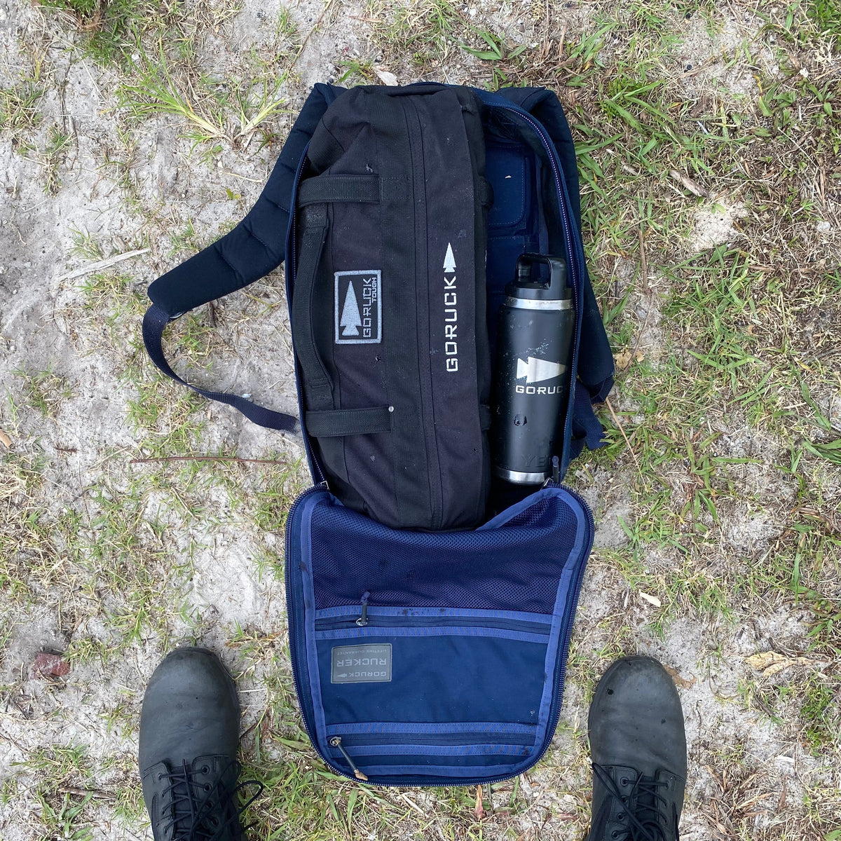 On grass, an open backpack reveals Simple Training Sandbags and a water bottle inside—ideal for sandbag workouts or your home gym. Black boots are visible below.