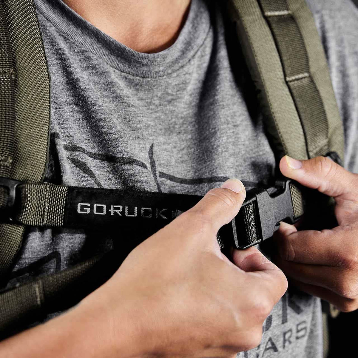 A person secures the Training Sternum Strap on an olive green GORUCK backpack with MOLLE webbing, while wearing a grey t-shirt.