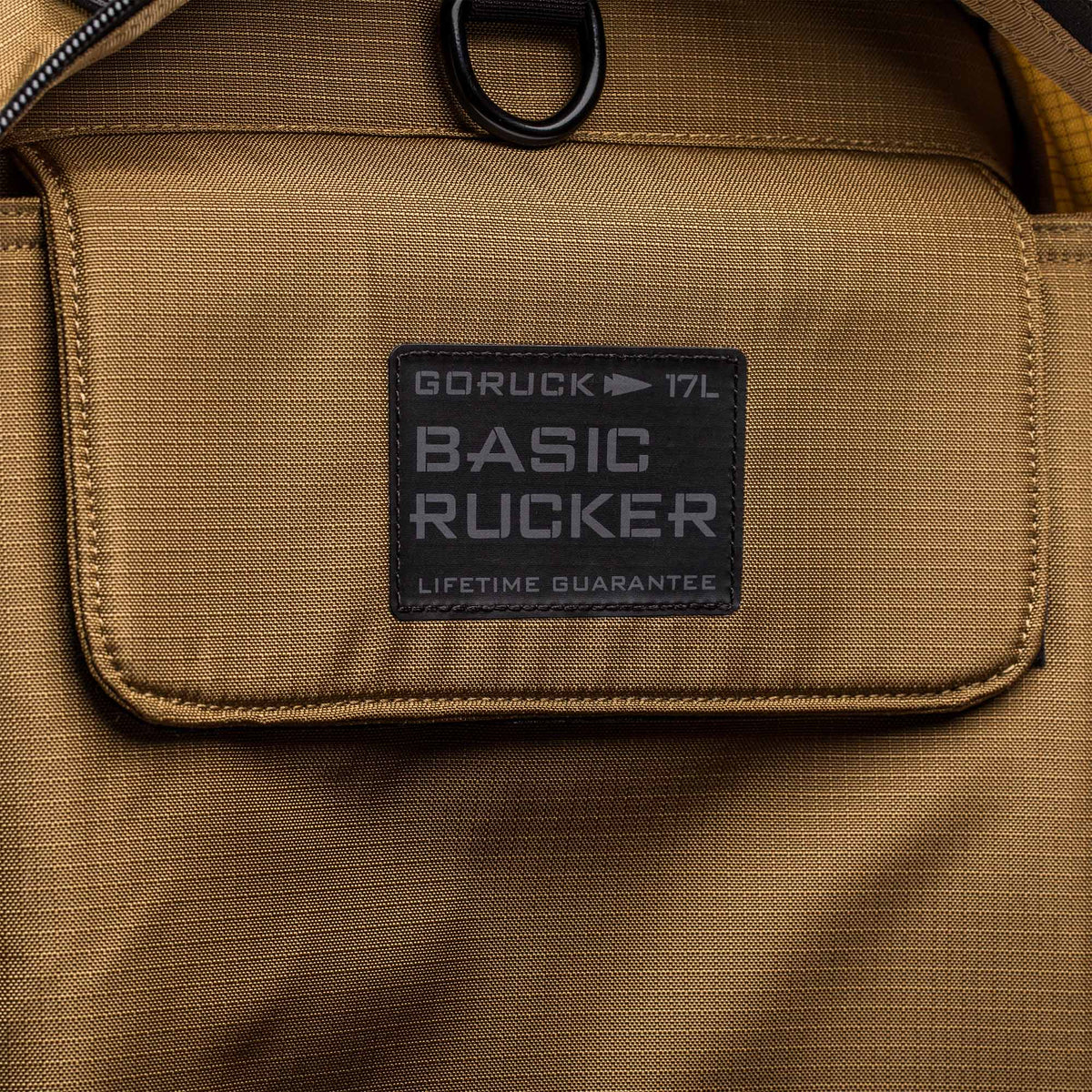 Close-up of a tan Basic Rucker® backpack pocket, featuring a label that reads 