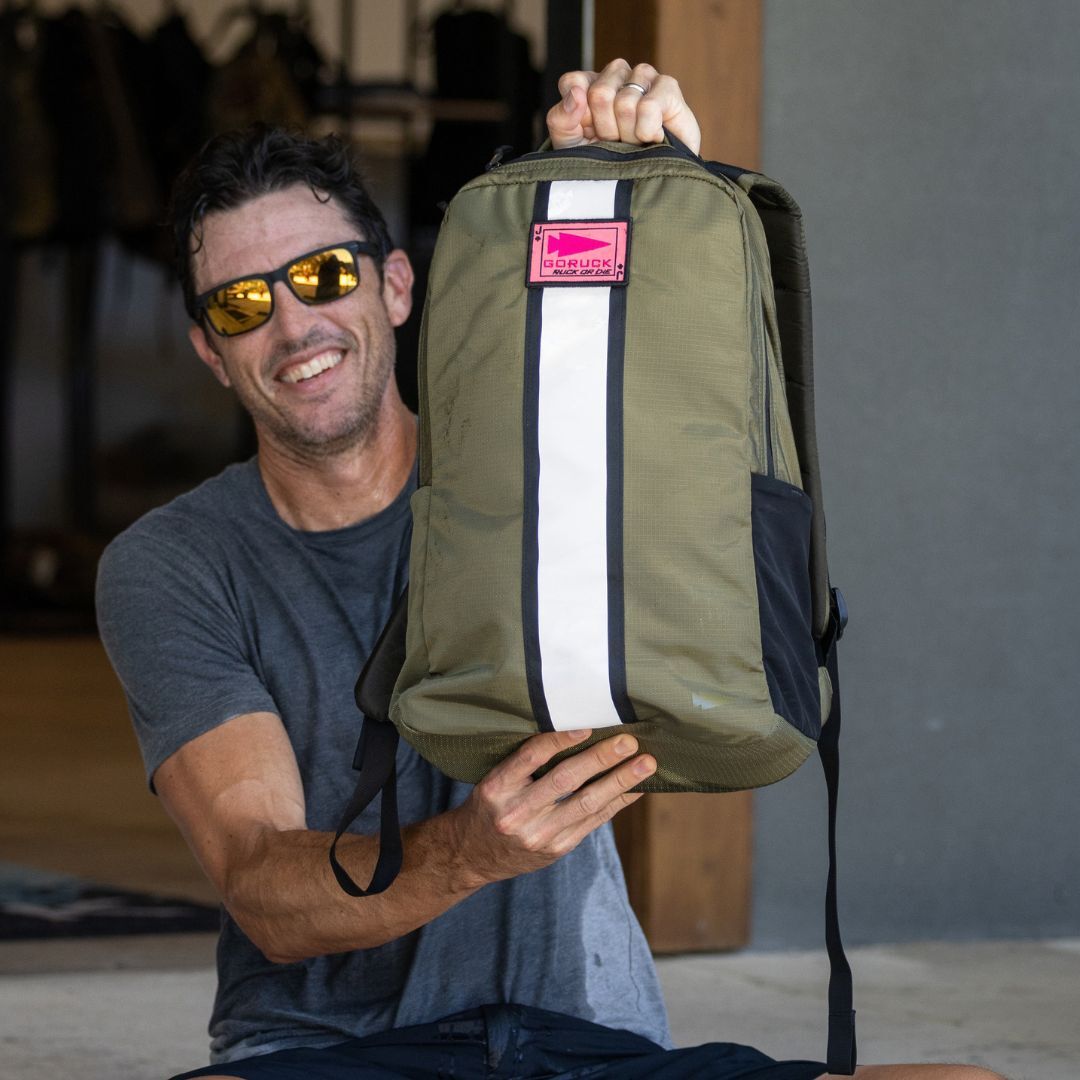 A smiling man wearing sunglasses holds up the Basic Rucker, a green rucksack featuring a white stripe and pink label.