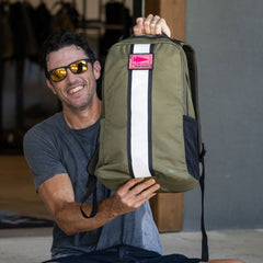 A smiling man wearing sunglasses holds up the Basic Rucker, a green rucksack featuring a white stripe and pink label.