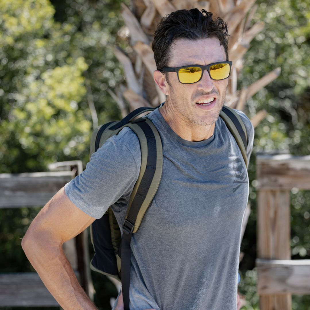 A man wearing sunglasses and a gray shirt stands outdoors with the Basic Rucker backpack, trees, and a fence behind him.