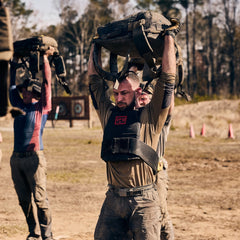 Three people lift heavy sandbags overhead while using the Rucker 5.0 during an outdoor rucking fitness challenge on a dirt field.