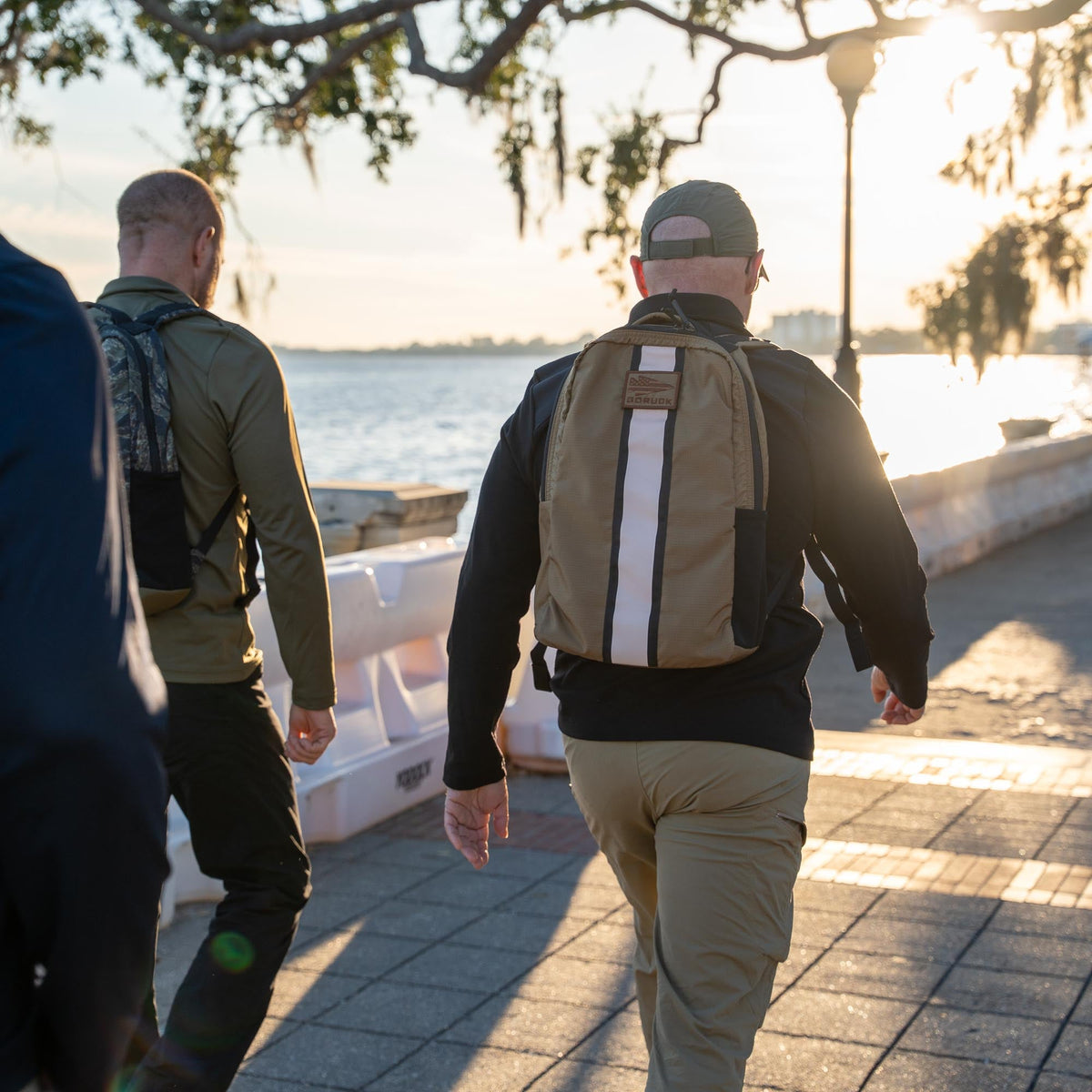 At sunrise, two men with backpacks stroll along a waterfront path, each carrying a Basic Rucker® as they take in the peaceful view.