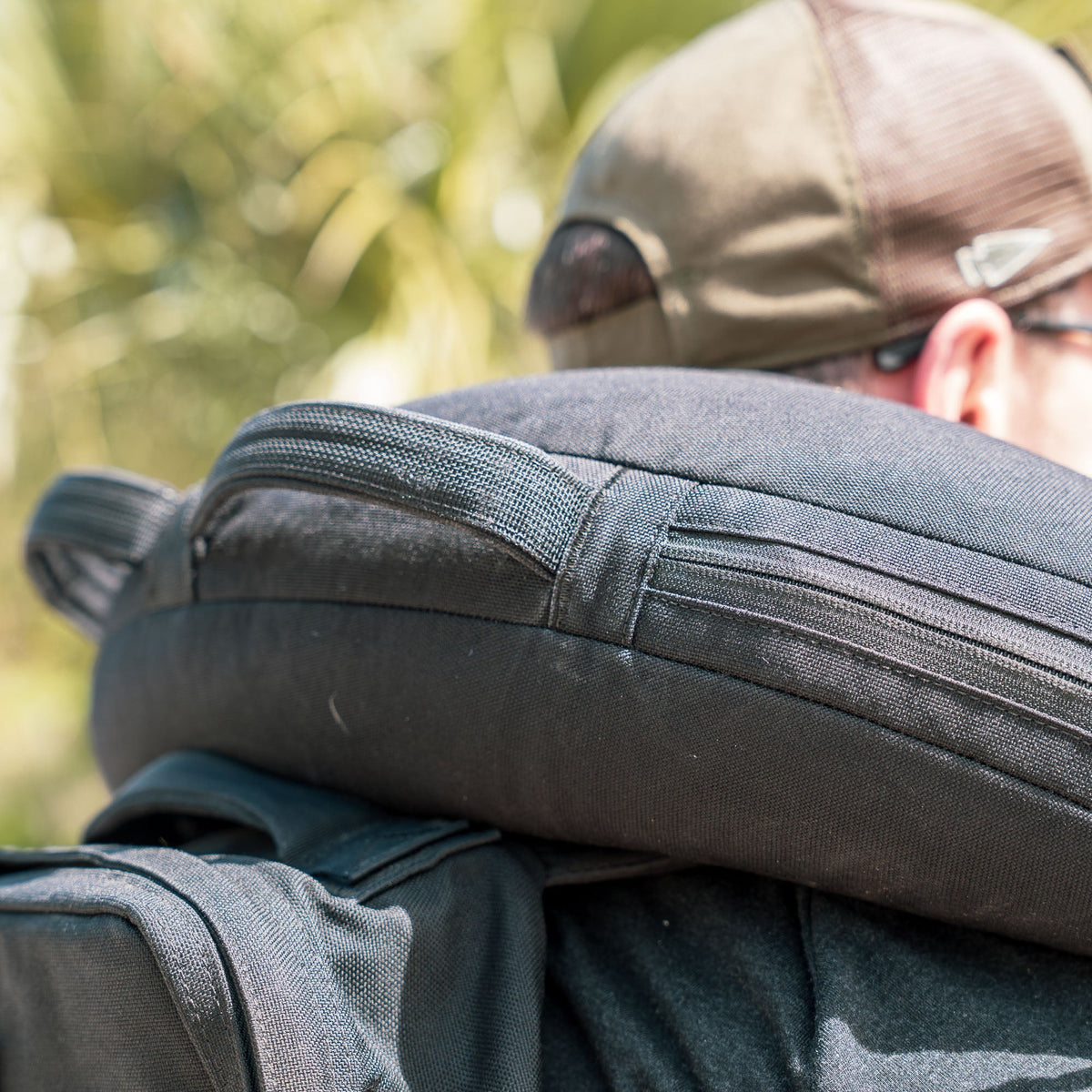 Close-up of a person outdoors with green foliage blurred in the background, carrying a dark backpack designed to hold a Bulgarian Sandbag.