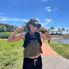 Smiling woman in sunglasses and a hat poses outdoors near the beach on a sunny day, wearing the Spy Ruck | Womens Weighted Vest.