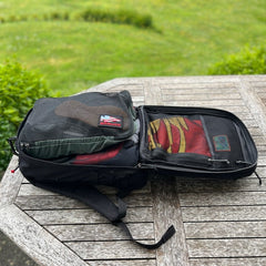 A GR2 ruck, opened on a wooden table outdoors amid greenery, features mesh compartments—ideal for one-bag travel and carry-on compliant adventures.