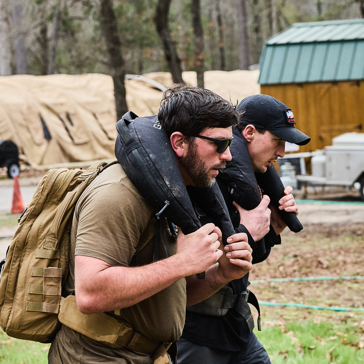 Two men outdoors carry Bulgarian Sandbags with reinforced handles on their shoulders during a fitness challenge.