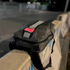 A Basic Rucker in gray, featuring a reflective stripe and a pink label, rests on a concrete barrier at night—ready for your next GORUCK rucking adventure.
