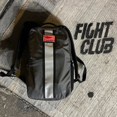 A Basic Rucker backpack in gray, featuring a red patch, rests on concrete beside the bold, black stenciled words “FIGHT CLUB,” embodying the rugged essence of rucking with this popular GORUCK product.