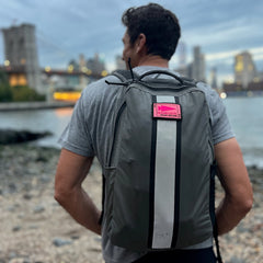 A man with short hair, wearing a gray shirt and carrying the Basic Rucker backpack, stands near a rocky waterfront with views of the city skyline.