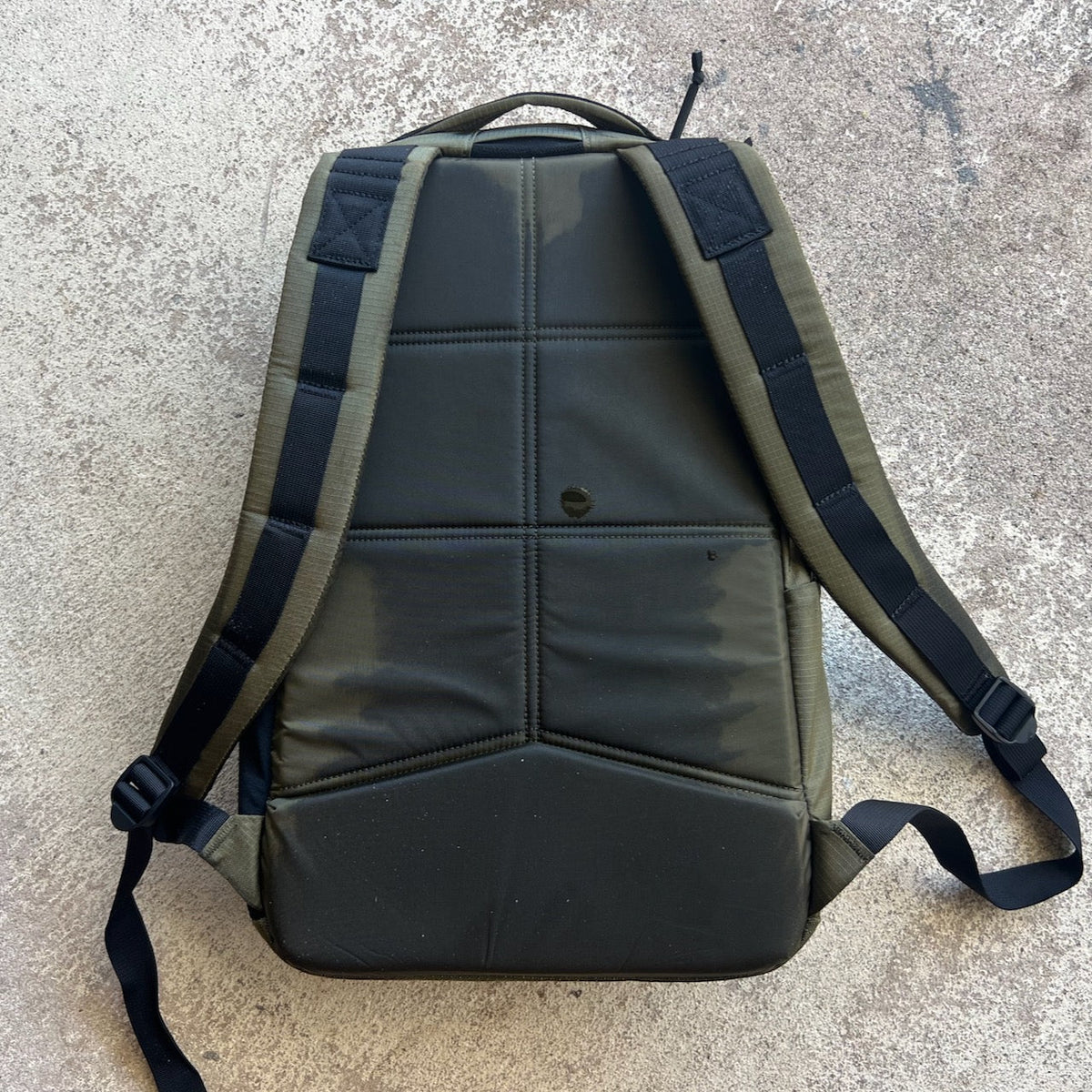 A Basic Rucker backpack in green with black straps lies flat on a textured, light-colored surface.