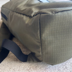 Close-up of the bottom corner of the Basic Rucker backpack in green with a black strap, resting on a light surface.