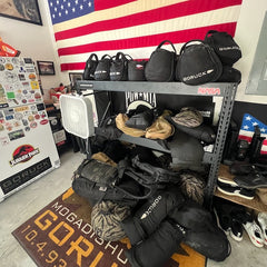 A shelf with GORUCK bags and gear, featuring Sand Kettlebells with padded handles, is set before a large American flag and a sticker-covered fridge.