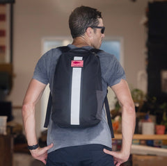 A man in sunglasses stands indoors, facing away from the camera with the Basic Rucker backpack in black.