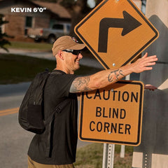 Man wearing a GORUCK backpack stands by caution blind corner sign, outdoors, 6 feet tall.