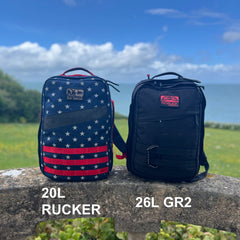 Two GR2 backpacks—one with an American flag pattern and one in black—sit on a stone ledge by the ocean, ideal for one bag travel adventures.