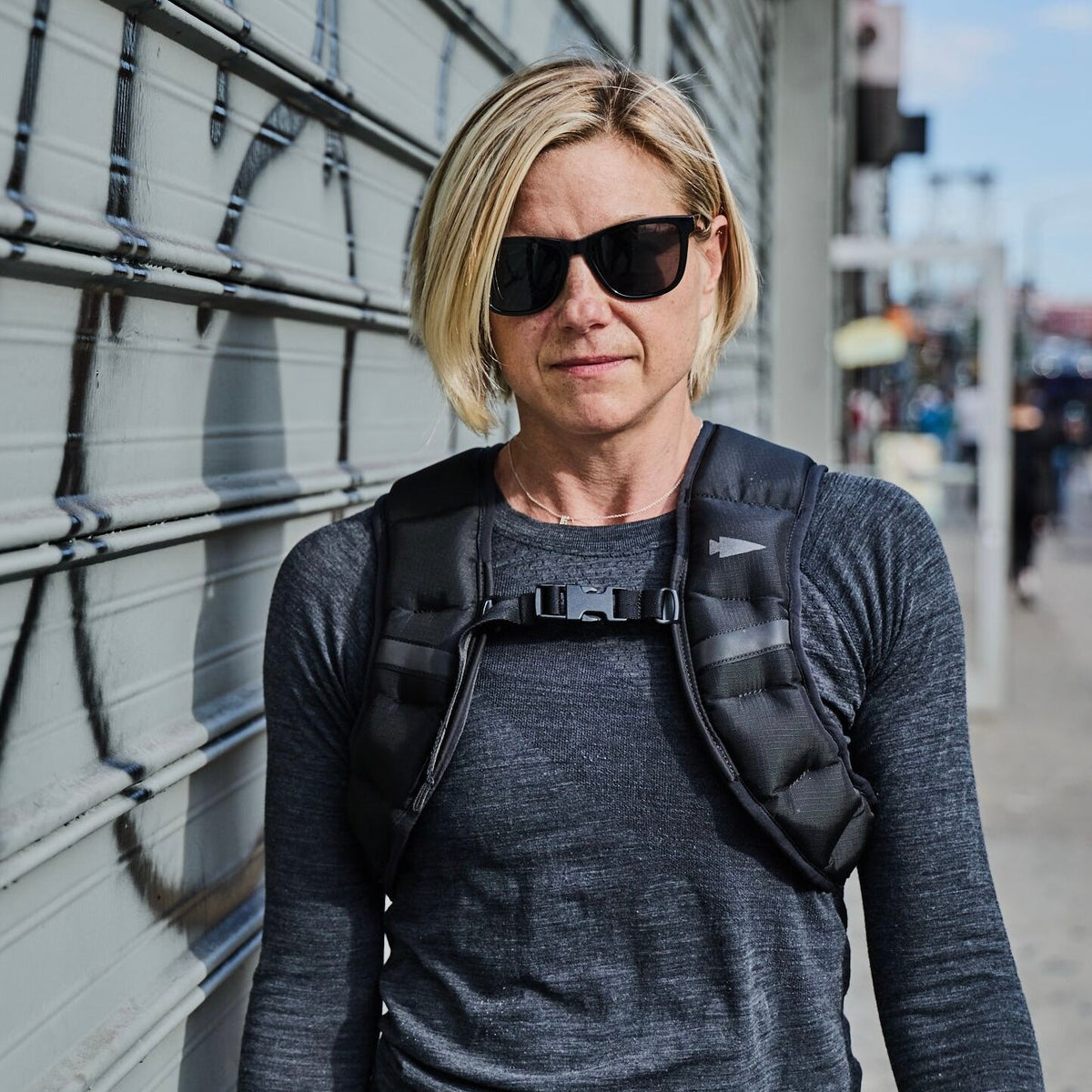 Blonde woman in sunglasses wearing a Spy Ruck | Women's Weighted Vest stands outdoors in front of a graffitied metal gate.