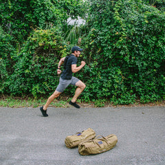 A man in athletic wear runs past two duffel bags on a paved path, wearing the Ruck Plate Carrier 3.0 for enhanced lumbar support, with dense greenery in the background.